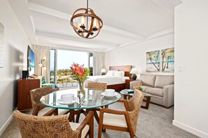 Ein Hotelzimmer mit einem Glastisch, Korbstühlen, einem Bett mit weißen Leinen, einer Couch und einem großen Fenster mit Blick auf Palmen und das Meer.