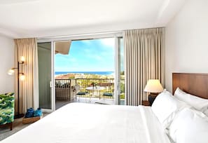 A hotel room with a bed, a nightstand, a lamp, a chair, a table, and a view of the ocean.