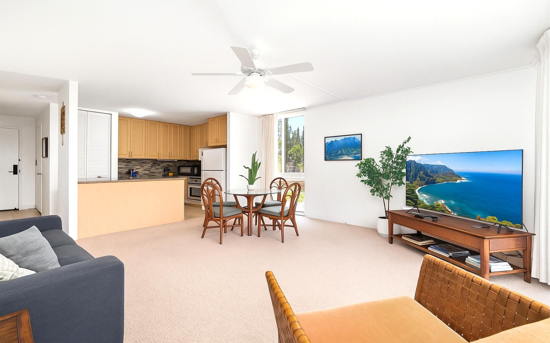 A modern living room with a sofa, a dining area with chairs, a kitchen with wooden cabinets, a ceiling fan, and a large TV displaying a scenic view.