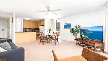 A modern living room with a sofa, a dining area with chairs, a kitchen with wooden cabinets, a ceiling fan, and a large TV displaying a scenic view.