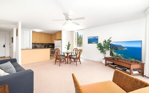 Een moderne woonkamer met een bank, een eethoek met stoelen, een keuken met houten kasten, een plafondventilator en een grote televisie waarop een mooi landschap te zien is.