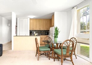 Une cuisine compacte avec des meubles en bois, un coin repas doté d’une table en verre et de chaises en osier, et une vue sur la verdure par une fenêtre.