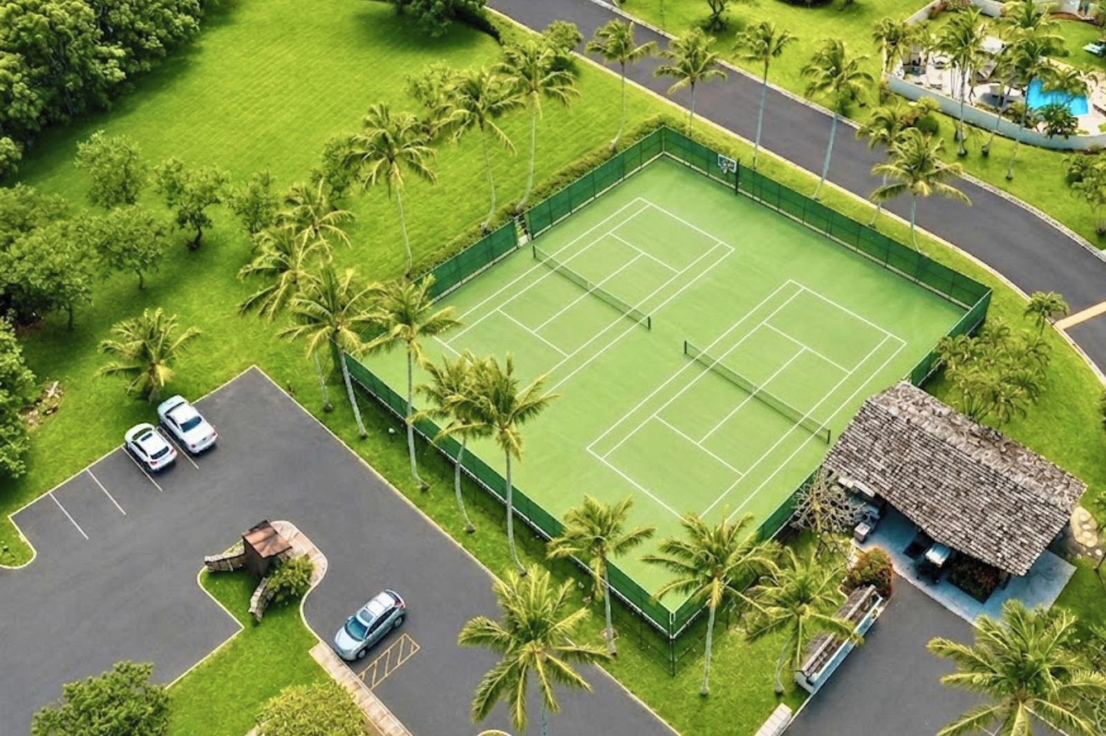 An aerial view of a tennis court surrounded by palm trees, with a parking lot and a building with a thatched roof.