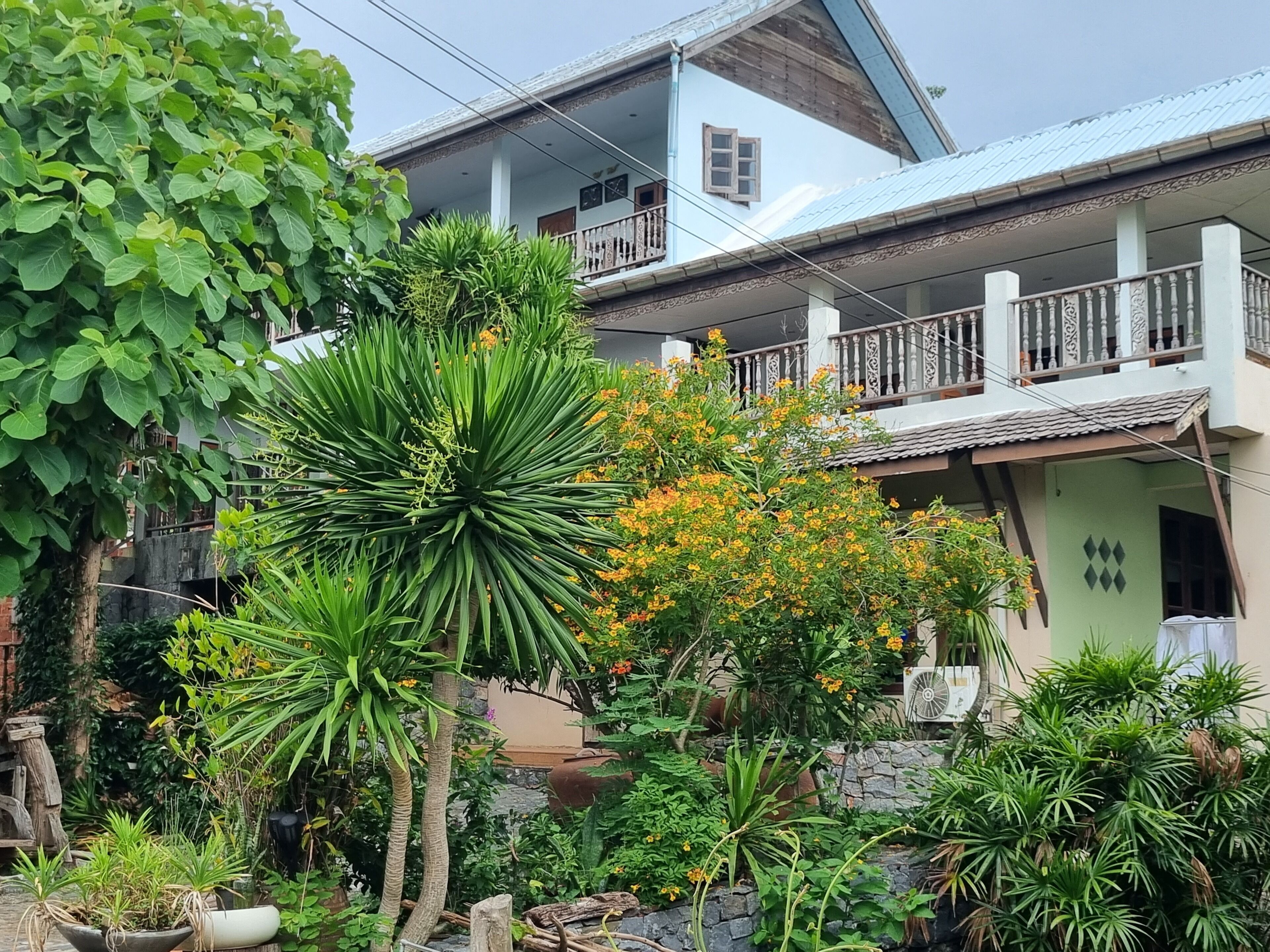 A two-story house with a balcony, surrounded by lush greenery and flowering plants.