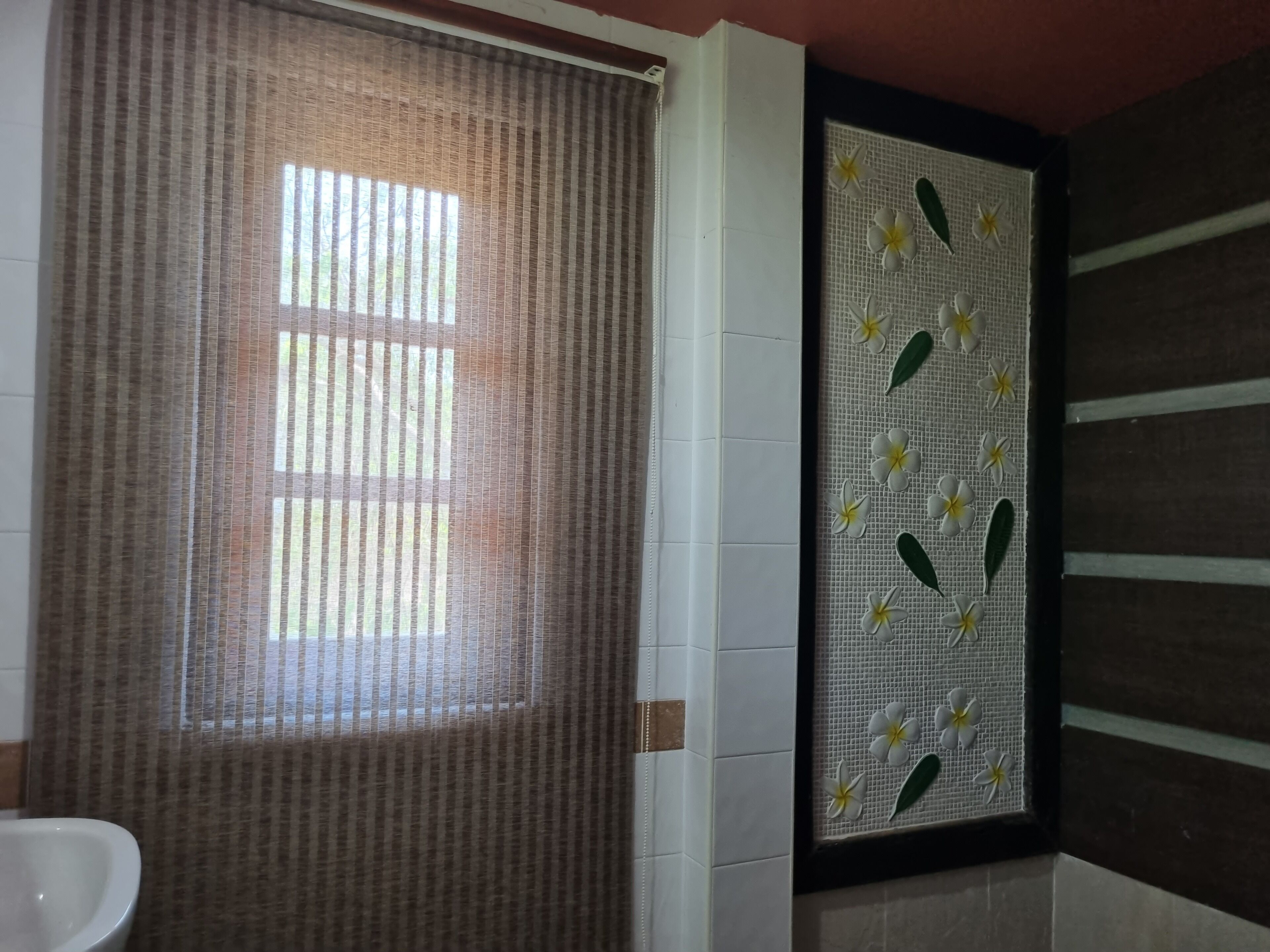 A bathroom with a window covered by a striped curtain, a white sink, and a decorative panel with flowers and leaves.