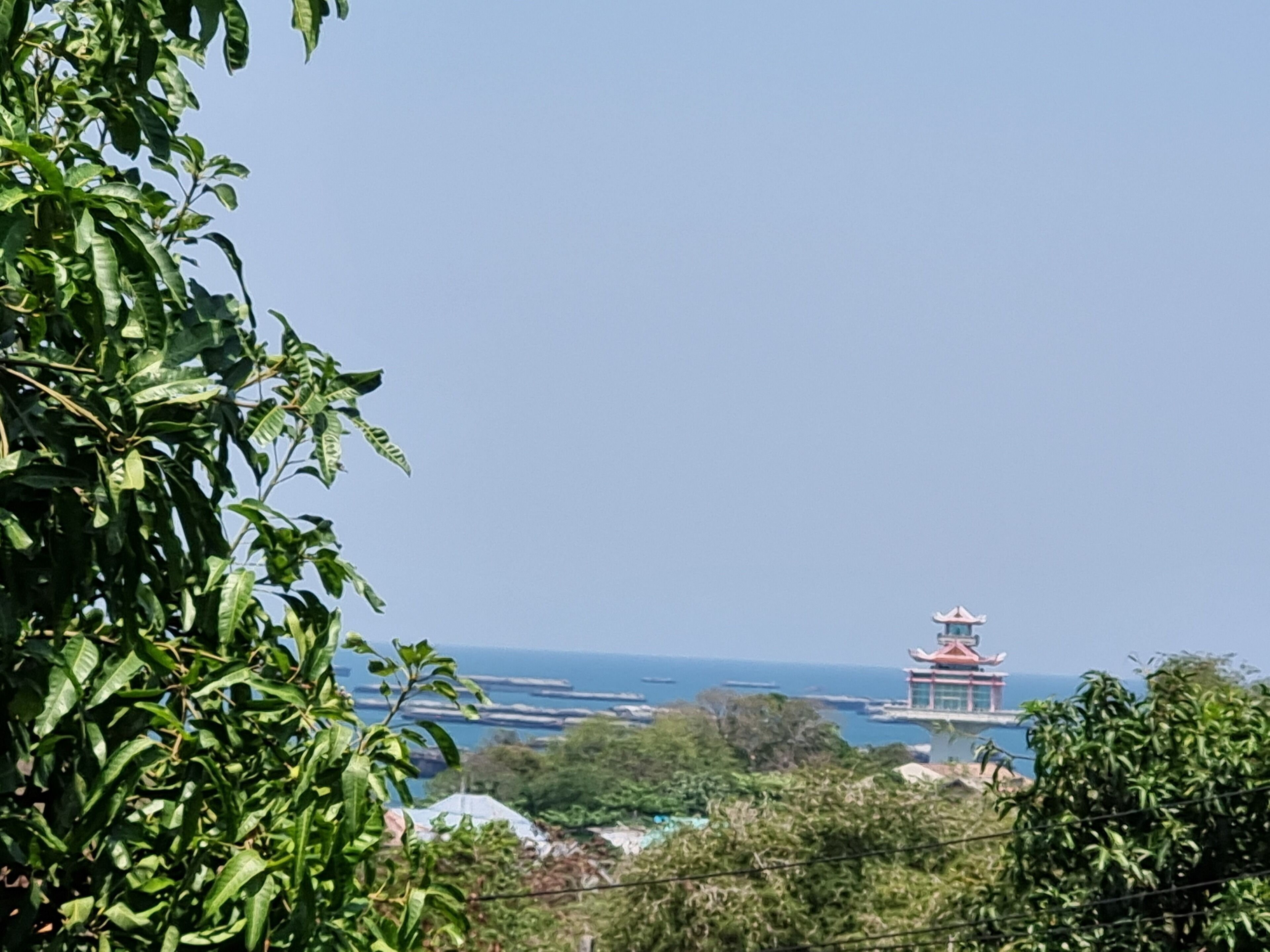 Vista costera con un edificio tradicional de techo rojo y paredes blancas, rodeado de abundante vegetación y con vista al mar.