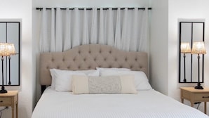 Un dormitorio con cabecera capitonada, una cama grande con ropa de cama blanca, dos mesitas de noche con lámparas y un banco al pie de la cama.