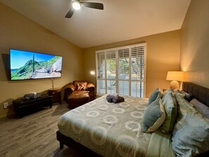 A bedroom with a bed, a TV, a chair, a window with blinds, and a ceiling fan.