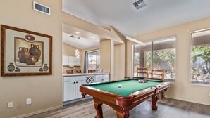 Uma mesa de bilhar em um espaço com cozinha, janelas amplas e um quadro na parede.
