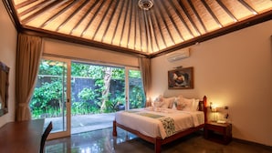 A bedroom with a large bed, a nightstand, a desk, and a view of greenery outside. - Luxury 3BR Kahuripan Villa (Bali)