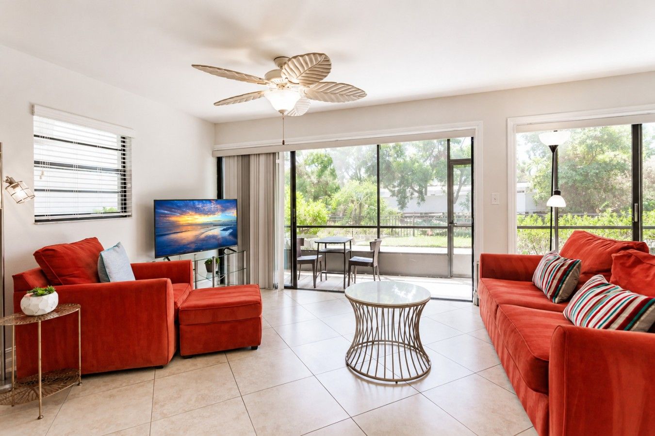 A modern living room with a red sofa set, a flat-screen TV, a ceiling fan, and large windows offering a view of greenery.