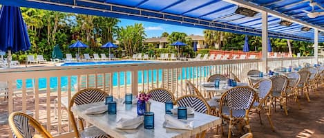 An outdoor pool area with lounge chairs and tables, a pool, and a covered seating area with umbrellas.