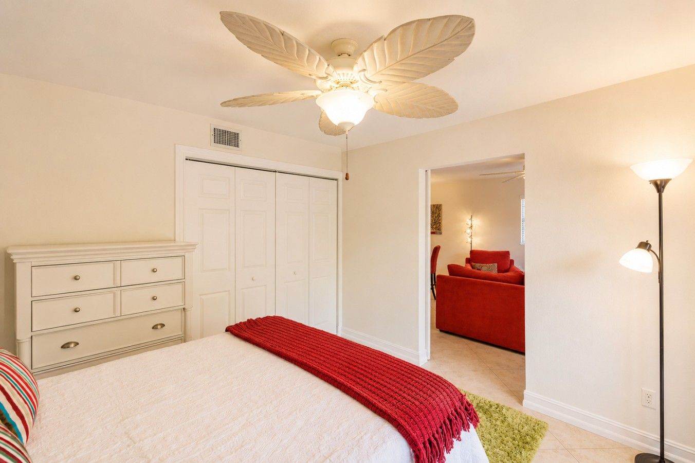 A bedroom with a bed, a dresser, a ceiling fan, a closet, and a red chair in the adjacent room.