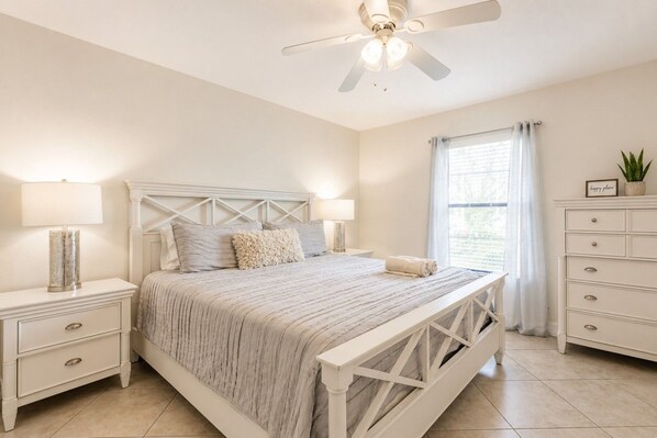 Un dormitorio con cama, mesitas de noche, cómoda, ventilador de techo y ventana con cortinas.