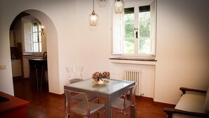 A dining area with a glass table and chairs, a window with a view of greenery, and a kitchen in the background. - Apartment in an old renovated mill with terrace (Capannori)
