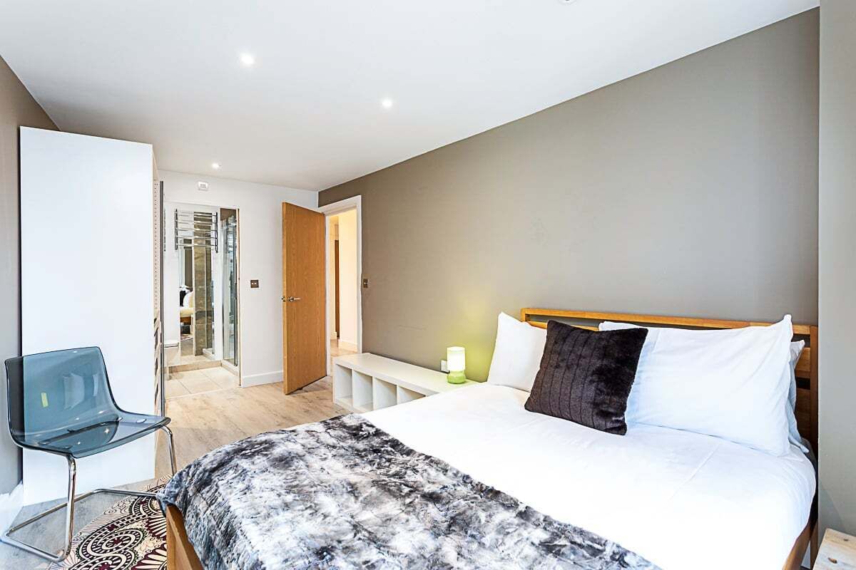 Une chambre d’hôtel moderne avec un grand lit, une table de chevet avec une lampe, une chaise en verre et une vue sur le couloir.