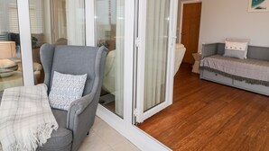 A living room with a grey sofa, a small table, and a bench with cushions. There is a sliding glass door leading to another room. - Haul a Gwynt (Holyhead)