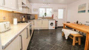 A modern kitchen with a built-in washing machine, a wooden dining table, and a bench. - Haul a Gwynt (Holyhead)