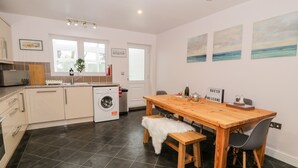 A modern kitchen with a washing machine, a wooden dining table, and a bench. There are three framed pictures on the wall. - Haul a Gwynt (Holyhead)