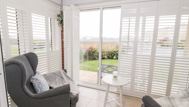 A room with a grey sofa, a small round table, and a view of a garden through large windows with white shutters. - Haul a Gwynt (Holyhead)