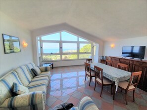 A spacious living area with a large window offering a view of the sea, a sofa, a dining table with chairs, and a flat-screen TV mounted on the wall. - “The Uncle Michele Penthouse” (Tropea)