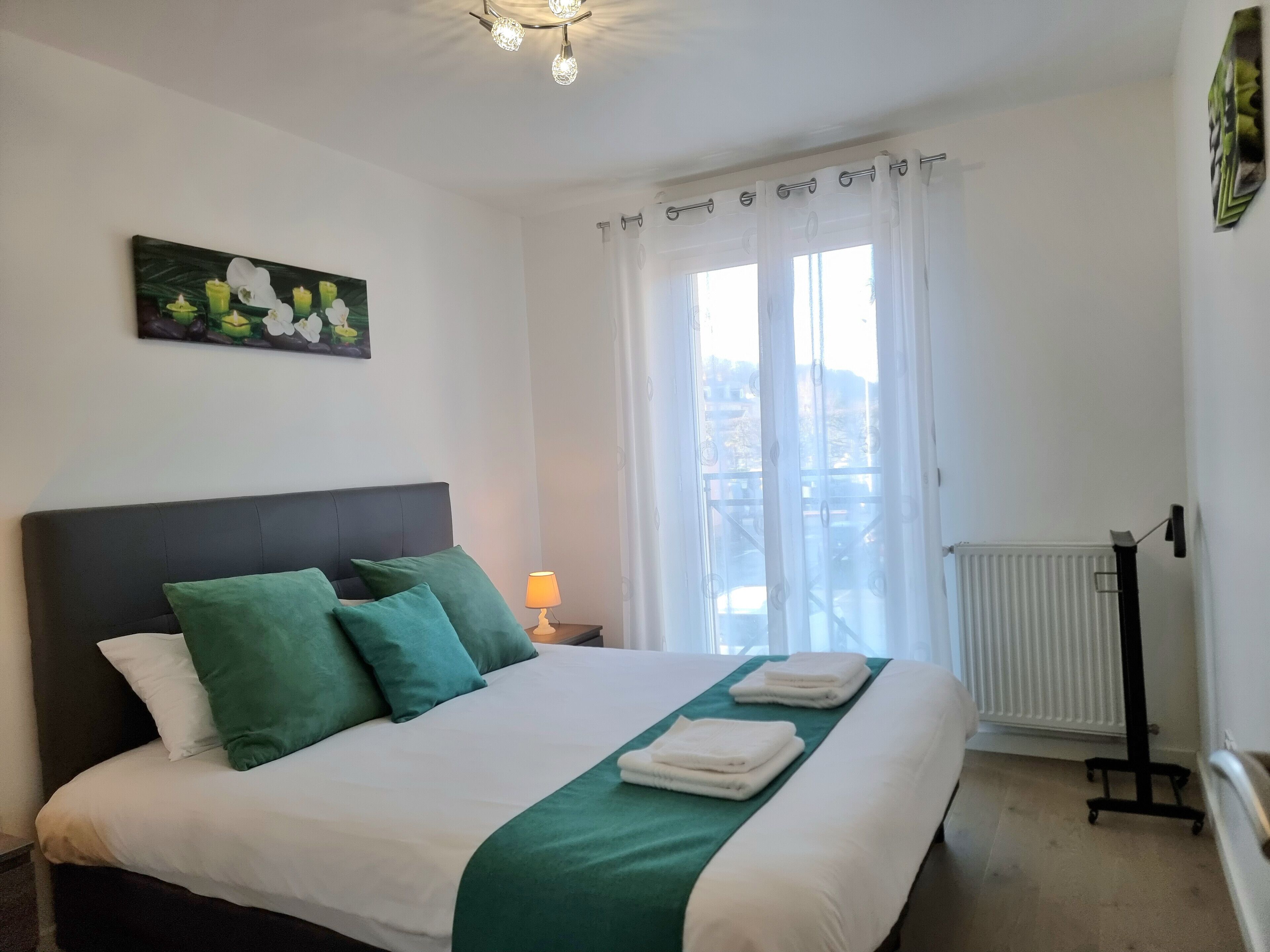 A neatly made bed with green and white bedding, a bedside lamp, and a view of the city through large windows.