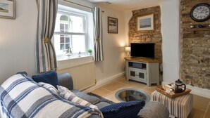 A cozy living room with a sofa, a television on a stand, a stone wall, and a window with curtains. - Honeymoon Cottage (Bideford)