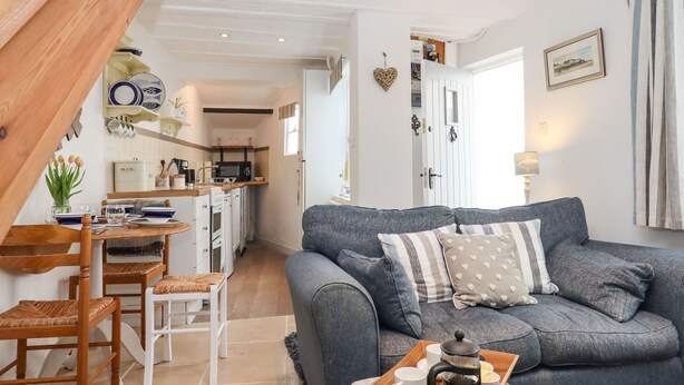 A cozy living room with a grey sofa, wooden dining table, and chairs. A kitchen is visible in the background with white cabinets and a microwave. - Honeymoon Cottage (Bideford)