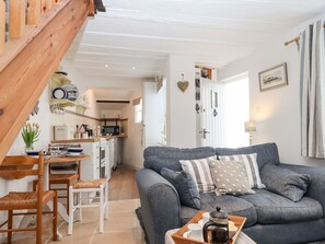 A cozy living room with a grey sofa, wooden dining table, and chairs. A kitchen is visible in the background with white cabinets and a microwave.