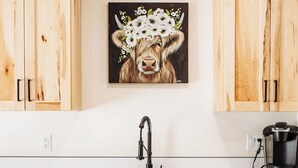 Une cuisine avec des meubles en bois, un lave-vaisselle en acier inoxydable, une machine à café noire et un tableau représentant une vache décorée de fleurs sur le mur.