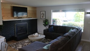A living room with a fireplace, a sofa, a TV mounted on a wooden wall, and a large window with a view of the outside.