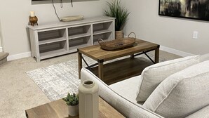 A living room with a flat-screen TV mounted on the wall, a white console table, a wooden coffee table, a white sofa, and a large abstract painting.