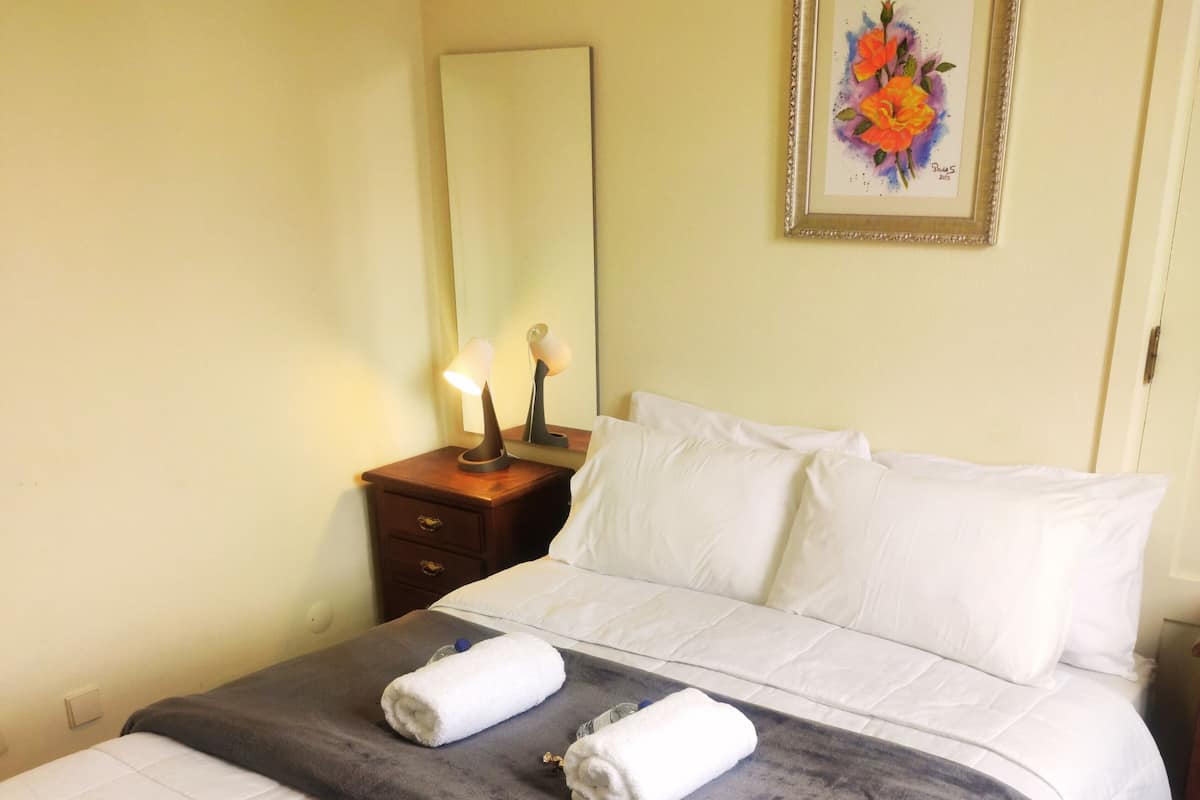 A neatly made bed with white linens and two rolled towels, a bedside table with a lamp, and a framed floral artwork on the wall.
