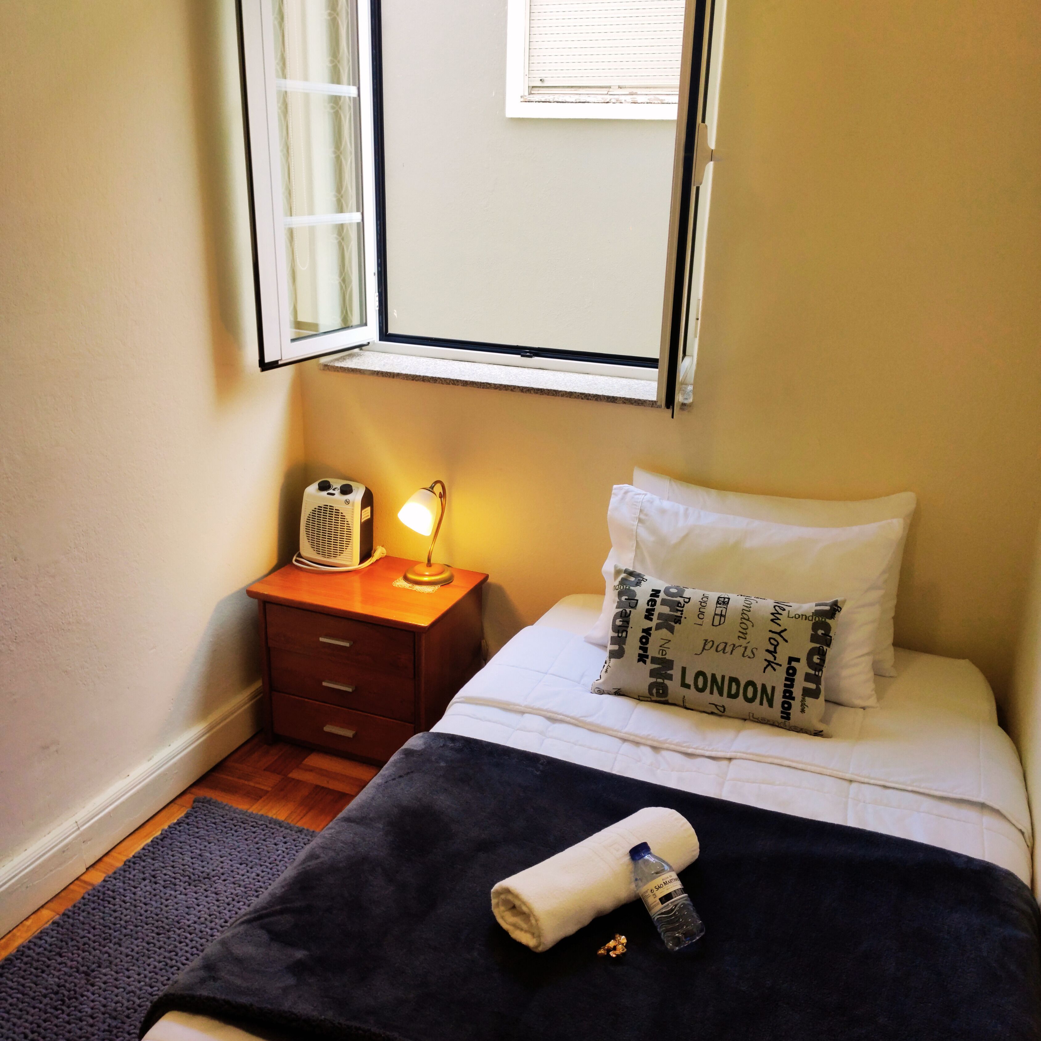 A bedroom with a bed, a bedside table with a lamp, a window, and a radiator.