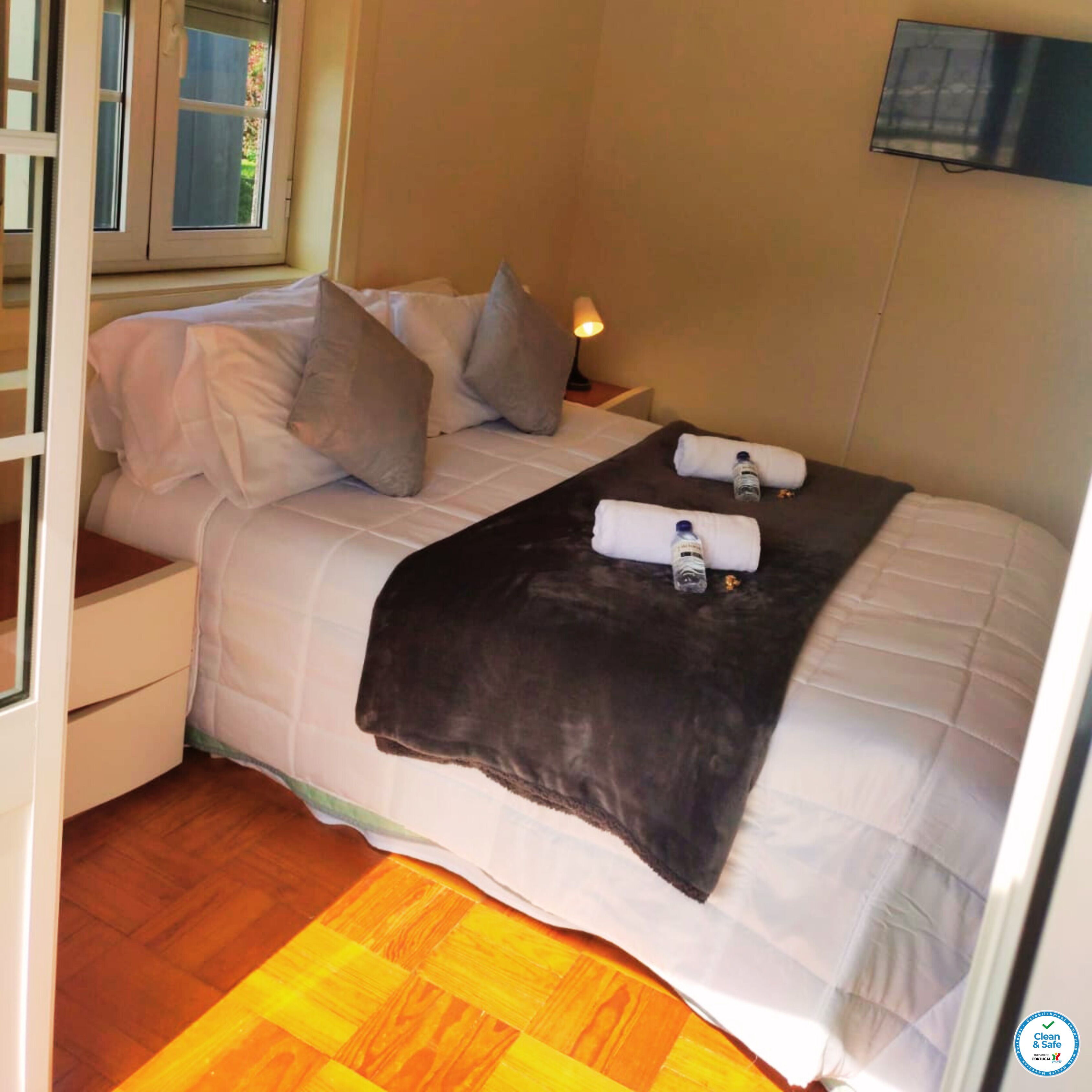 Cama arrumada com almofadas, manta e duas toalhas com garrafas. Criado-mudo com abajur e janela com cortinas.