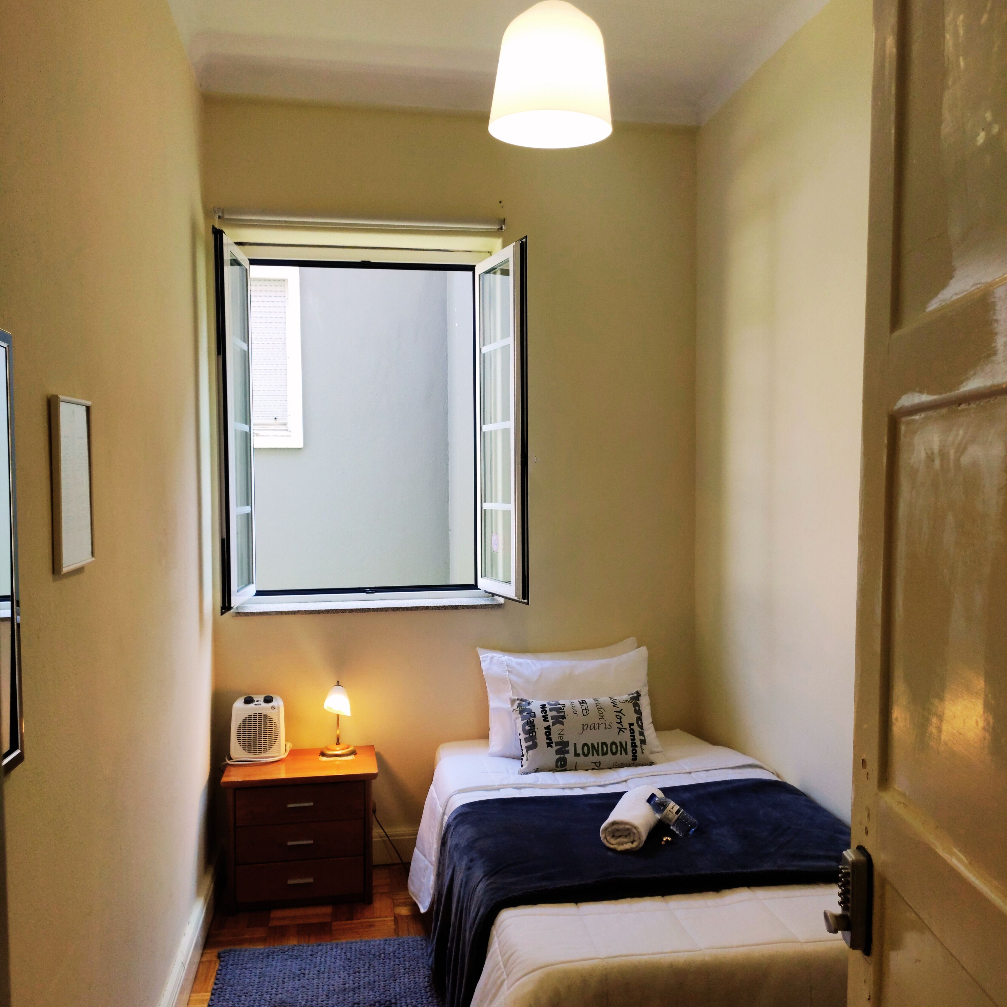 A small, narrow hotel room with a single bed, a bedside table with a lamp, a window with blinds, and a door.