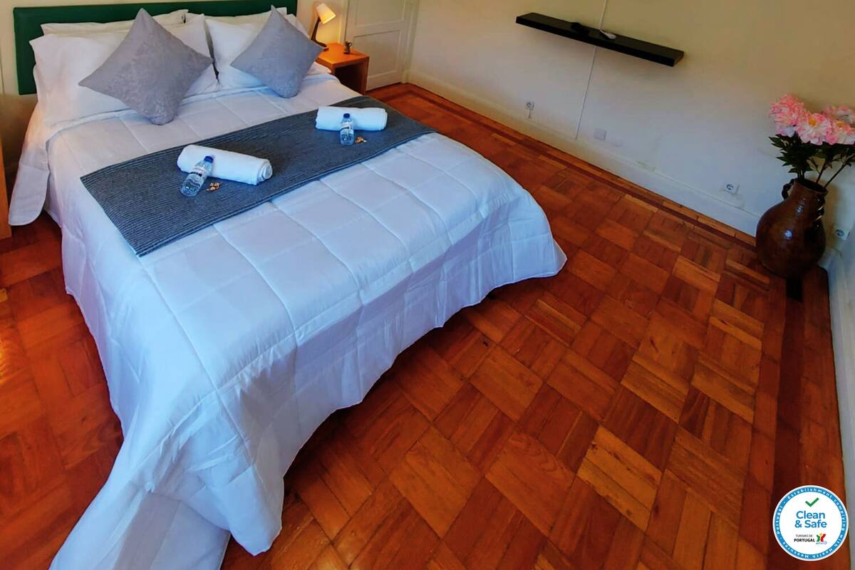 A neatly made bed with pillows and a folded towel, a bedside table with a lamp, a flat-screen TV mounted on the wall, and a wooden floor.