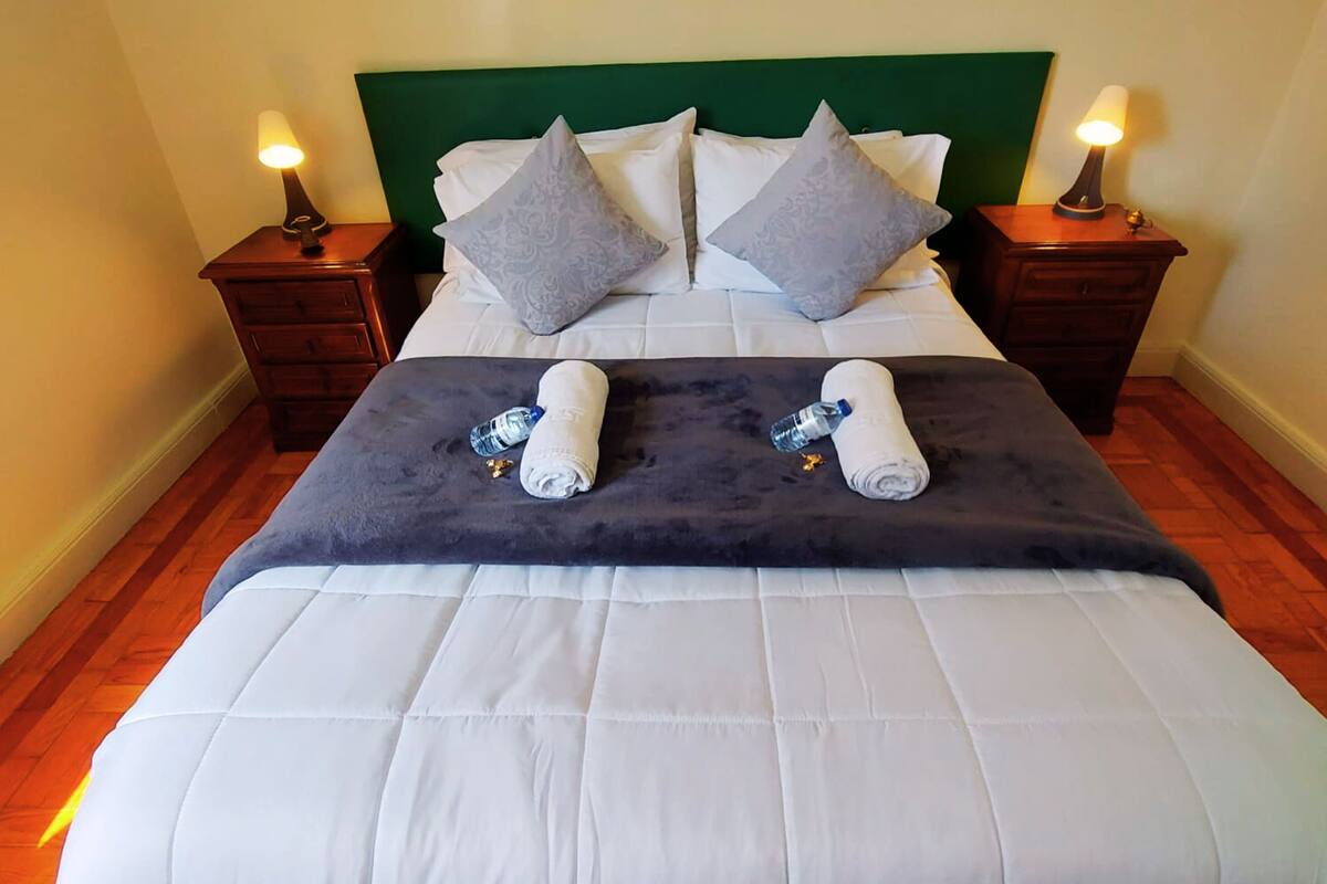 A neatly made bed with white and grey bedding, two rolled towels, and a water bottle on a dark blue blanket.