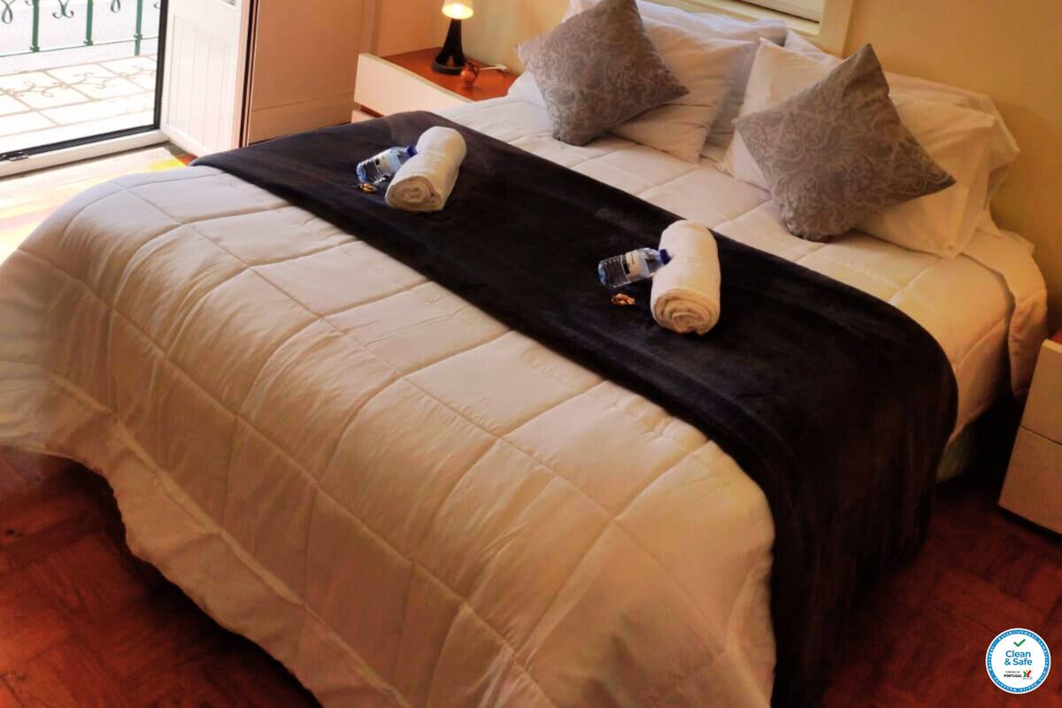 A hotel room with a bed, pillows, a nightstand with a lamp, and a window with blinds.