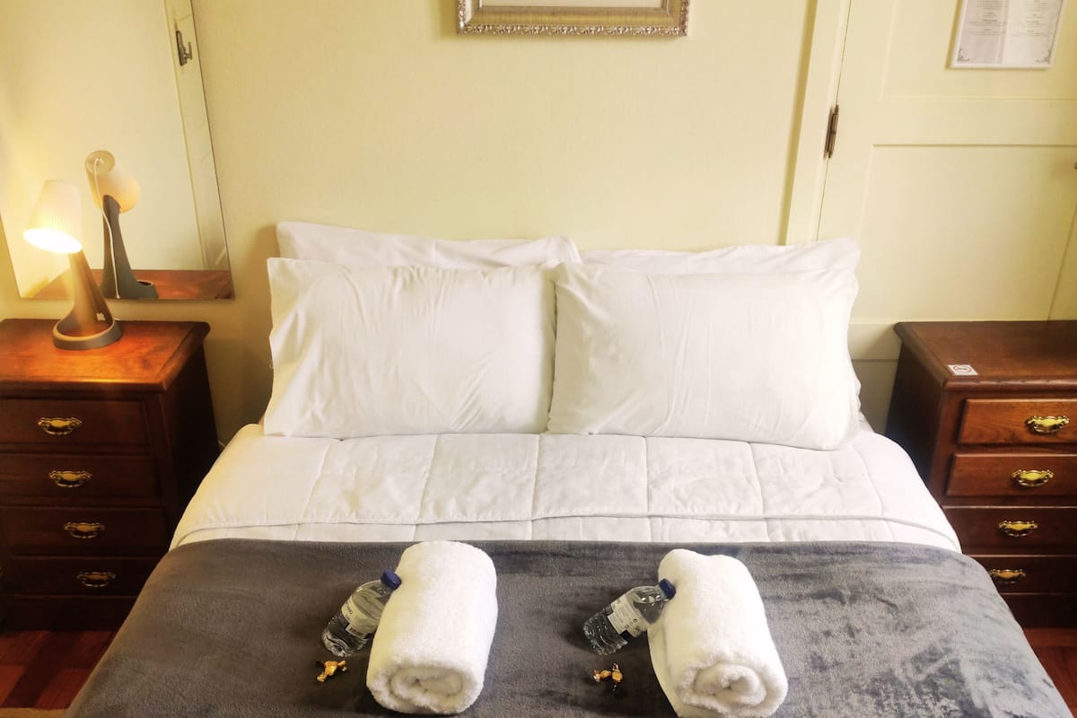 A hotel room with a bed, two pillows, a blanket, two towels, a water bottle, and a lamp on a nightstand.