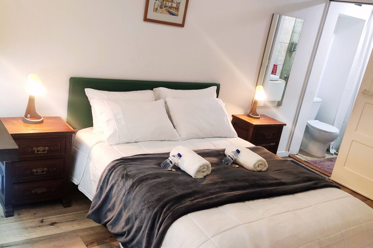 A neatly made bed with white linens, a dark blanket, and two rolled towels. There are two bedside lamps, a wooden nightstand, and a framed picture on the wall.