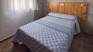 Een slaapkamer met een bed, een houten hoofdbord, een vloer met een stenen patroon, een raam met dunne gordijnen en een schilderij aan de muur.
