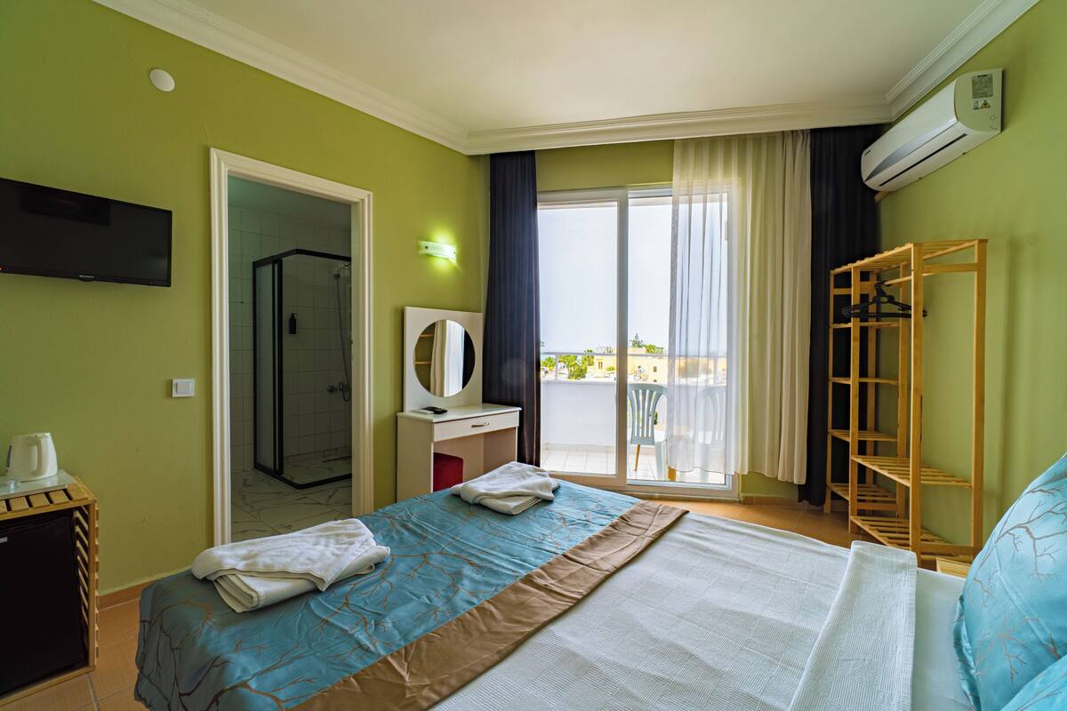 Een hotelkamer met een bed, een bureau, een spiegel, een televisie, een balkon met een stoel, en een airconditioning.