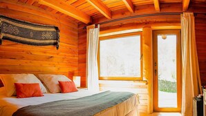 Un dormitorio de cabaña de madera acogedor, con cama, almohadas, mesita de noche, lámpara y ventana con cortinas.