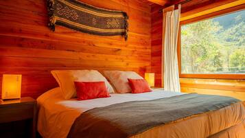 A wooden cabin bedroom with a bed, pillows, a blanket, a nightstand, a lamp, and a window with a view of trees.