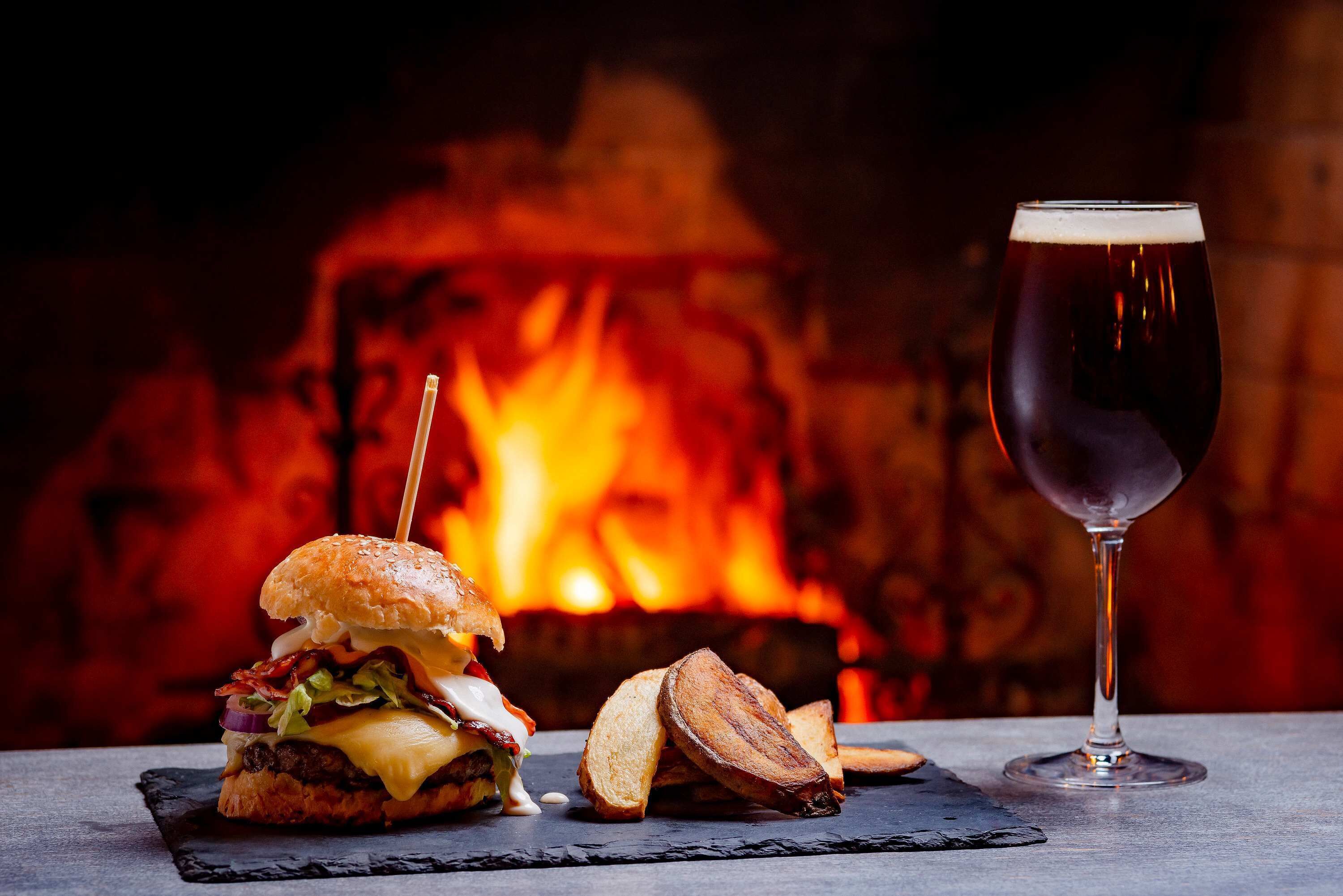 A burger with a toothpick, a glass of beer, and toasted bread on a slate plate, with a fireplace in the background.
