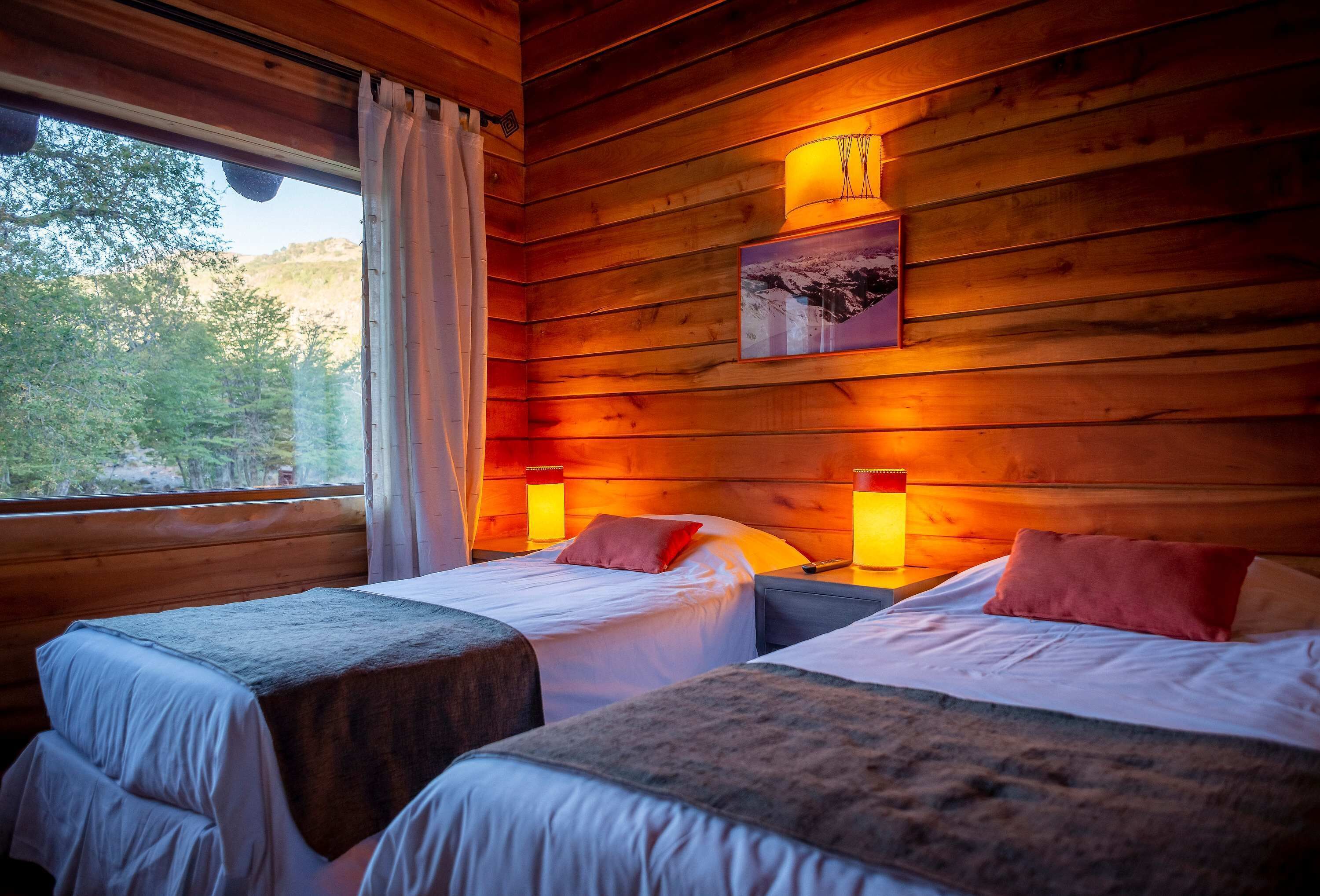 A cabin with two beds, wooden walls, a window with curtains, and a wall-mounted lamp.