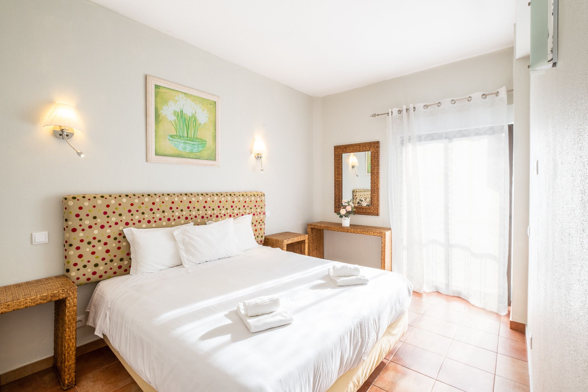 Ein Hotelzimmer mit einem Bett, zwei Nachttischen, einer Wandleuchte, einem Gemälde, einem Spiegel und einem fenster mit Vorhängen.
