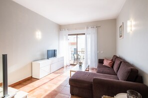 A modern living room with a sofa, a TV on a white cabinet, and a balcony with a view. - #099 T1 Caliços by Home Holidays (Albufeira)