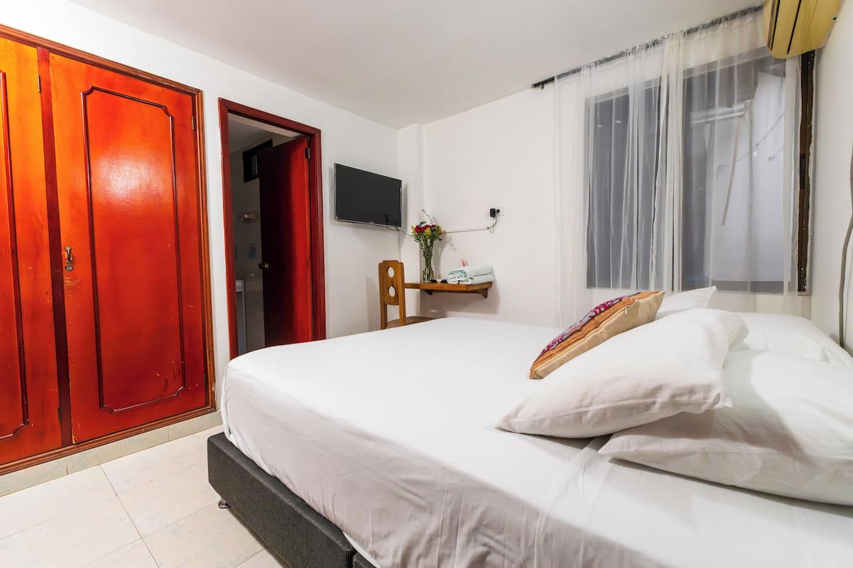Une chambre d’hôtel avec un lit, un téléviseur fixé au mur, une armoire en bois et une fenêtre avec des rideaux légers.